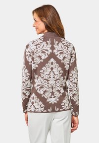 Pull marron à motifs avec un motif floral blanc, tissu texturé, col rond et manches longues, porté avec un pantalon de couleur claire.