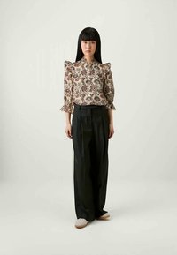Floral print blouse with puffed sleeves, buttoned front, paired with high-waisted black trousers and beige shoes, standing against a light backdrop.