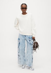 Γυναίκα που φοράει λευκό φαρδύ φούτερ, ανοιχτό μπλε παντελόνι καμπάνα cargo, ασημί αθλητικά παπούτσια, σκοτεινά γυαλιά ηλίου, κρατώντας καφέ τσάντα.