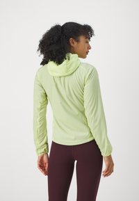 Giacca leggera verde chiaro con cappuccio e maniche lunghe, caratterizzata da una texture liscia e un design aderente; abbinata a leggings burgundy scuri.