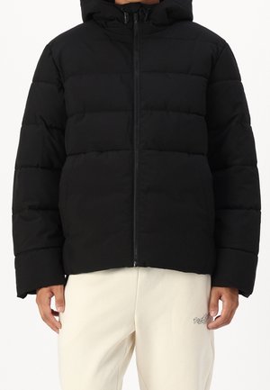 Osoba nosící černou prošívanou bundu se zapínáním na zip vpředu a kapucí, v kombinaci se světle béžovými kalhotami s malým textem na horní části stehna.