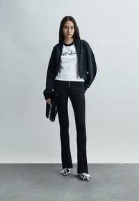 Femme debout contre un mur gris, portant une veste noire zippée, un T-shirt blanc avec logo, un pantalon noir, une pochette à franges et des chaussures métalliques pointues.