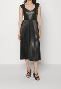 Robe midi plissée noire en cuir avec un corsage ajusté et des manches courtes ; associée à des sandales à talons hauts noires.