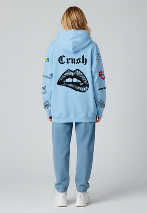 Felpa azzurra con grafica di labbra nere e testo "Crush", con ulteriori stampe sulle maniche; abbinata a pantaloni jogger blu coordinati.