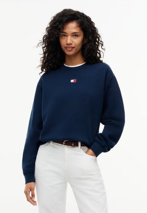 Kvinde med krøllet hår iført en mørkeblå sweatshirt med et lille logo og hvide bukser, stående med den ene hånd i lommen.