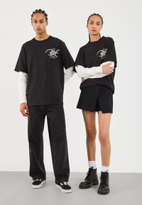 Storede oversized T-shirts i sort med grafiske designs, parret med denim og skræddersyede shorts. Fodtøj inkluderer sorte støvler og sneakers.
