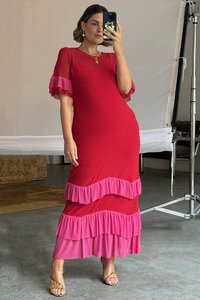 Röd maxiklänning med rosa volangdetaljer vid ärmarna och fållen. Tyget verkar slätt och lätt. Bärs med remsandaler.