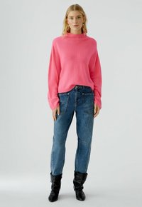 Rosa Strickpullover mit hohem Kragen, weiten Ärmeln und lockerer Passform, kombiniert mit blauen Jeans mit aufgesetzten Taschen und einem ausgeblichenen Look.
