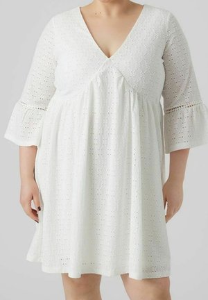 Person i plusstørrelse iført en hvid kjole af øjet stof med 3/4 klokkeærmer og V-udskæring, stående mod en ensfarvet lys grå baggrund.