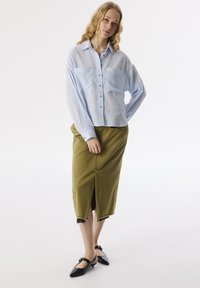 Camisa de botones azul claro con bolsillos frontales, combinada con una falda verde olivo hasta la rodilla con una abertura frontal. Bailarinas negras de punta.