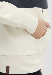 Sweat-shirt avec une partie supérieure bleu marine, un corps crème et une poche avant dissimulée ; fini avec une étiquette de marque marron sur l'ourlet inférieur.