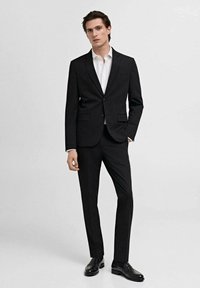 Zwart op maat gemaakt pak met gekartelde revers, aansluitende blazer en broek, gecombineerd met een wit overhemd en zwarte leren schoenen.