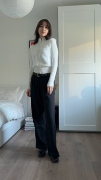 Gilet blanc duveteux porté sur un haut à manches longues côtelé, associé à un pantalon noir à jambes larges. Le décor comprend un lit blanc et un sol en bois clair.