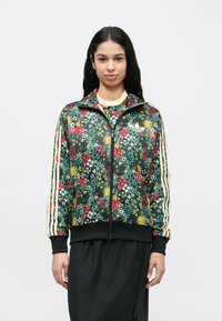 Frau mit langen schwarzen Haaren trägt eine bunte, mit Blumenmuster versehene Zip-up-Jacke mit gelb gestreiften Ärmeln und einen schwarzen Rock vor einem schlichten weißen Hintergrund.