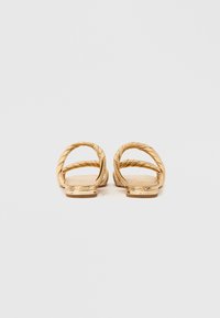 Χρυσά σανδάλια τύπου σλάιντ με στριφογυριστούς ιμάντες, επίπεδη σόλα και λεία υφή. Μινιμαλιστικός σχεδιασμός, με μεταλλική λεπτομέρεια στη φτέρνα.