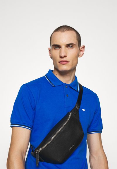 Μαυρη δερμάτινη τσάντα σλάινγκ με γυαλιστερή επιφάνεια και ασημένιο φερμουάρ, φορεμένη διαγώνια στο σώμα. Διαθέτει ιμάντα ώμου και διακριτική σήμανση.