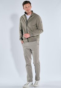 Lightweight olive green bomber jacket with a high collar, zipper closure, and ribbed cuffs, paired with light gray trousers and white sneakers.