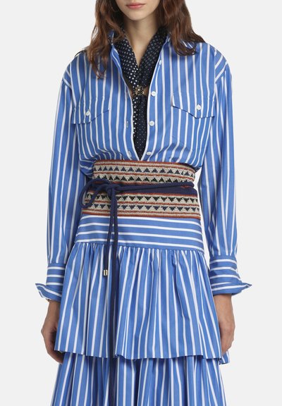 Femme portant une robe chemise à rayures verticales bleu et blanc avec une large ceinture à motifs et un foulard noir à pois noué autour du cou.