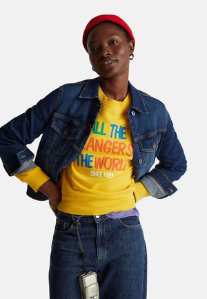 Persona con berretto rosso, felpa gialla con testo colorato, giacca di jeans e jeans con una fotocamera argento appesa all'asola della cintura.