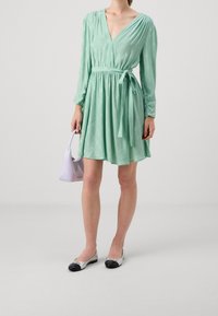 Vrouw in mintgroene wikkeljurk met een tailleband, houdt een kleine lila handtas vast en draagt zilveren flats met zwarte neuskapjes.