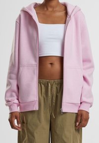 Sudadera con cremallera rosa en tejido suave con un ajuste holgado y bolsillos delanteros grandes, combinada con un top corto blanco y pantalones beige.