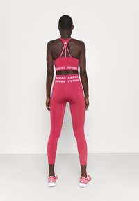 Rosa träningsoutfit bestående av en sport-bh med korsade axelband och högmidjade leggings, båda prydda med iögonfallande vit Adidas-logotyp.