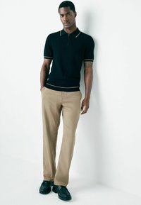 Man wearing black short-sleeve polo, beige pants, and black shoes stands relaxed against white wall with hands partially in pockets.