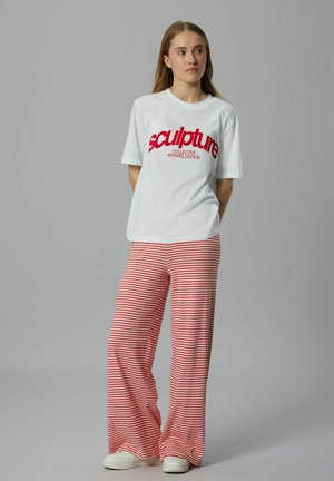Jonge vrouw staat met haar handen achter haar rug, draagt een wit T-shirt met rode tekst en rood-witte gestreepte wijde broek en witte schoenen.