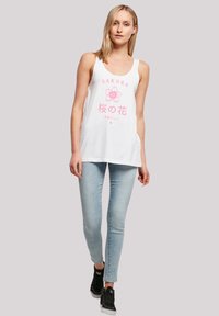 White tank top featuring pink "SAKURA" text and flower graphic. Paired with light blue skinny jeans and black sneakers. Smooth fabric.