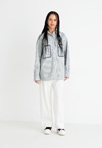 Camicia azzurro chiaro con bottoni, due grandi tasche e righe verticali, abbinata a pantaloni bianchi a gamba larga e scarpe chiare.