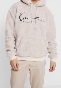 Beige Fleece-Pullover mit Kapuze, Fronttasche, Kordelzug und schwarzem gesticktem Logo. Weiche Textur und gerippte Bündchen.