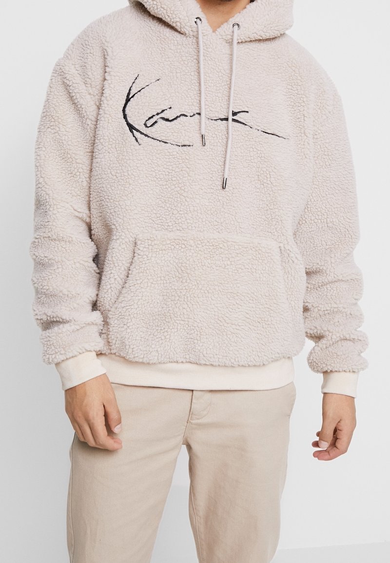 Beige Fleece-Pullover mit Kapuze, Fronttasche, Kordelzug und schwarzem gesticktem Logo. Weiche Textur und gerippte Bündchen.