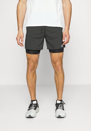 Man draagt zwarte gelaagde sportkorte broek, witte en zwarte hardloopschoenen en een wit sportshirt van mesh, staand tegen een effen achtergrond.