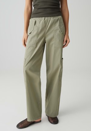 Personne portant un pantalon cargo vert olive avec de grandes poches frontales, associé à des chaussures à enfiler en maille noire, debout sur un fond uni.