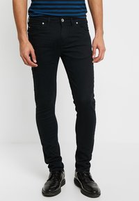 Vaqueros negros de corte ajustado con diseño de cinco bolsillos, hechos de denim, con cierre estándar de botón y cremallera, combinados con zapatos de vestir negros.