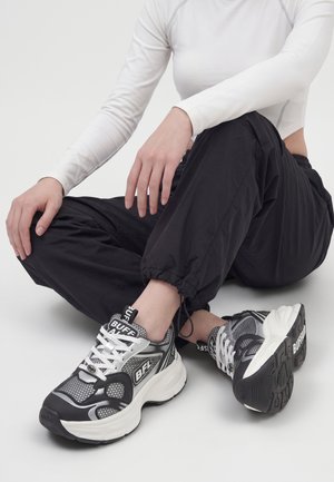 Person, der sidder med benene krydsede iført sort-hvide chunky sneakers med "BUFF" mærke og sorte bukser med elastiske manchetter.