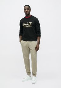 Čierna mikina s kapucňou so zlatým logom "EA7 Emporio Armani", v kombinácii s béžovými teplákmi a bielymi teniskami, na jednoduchom bielom pozadí.