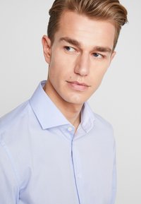 Camicia azzurra chiara con colletto rigido, chiusura a bottoni e delicato motivo a righe all'interno del colletto; tessuto di cotone liscio.