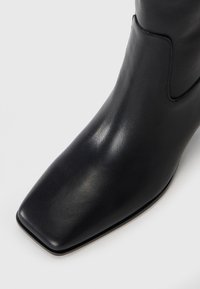 Stivale alla caviglia in pelle nera con punta quadrata e texture liscia. Presenta un design semplice con accenti minimi e una silhouette elegante.