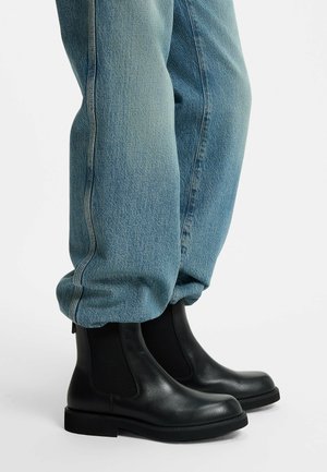 Gambe che indossano jeans blu larghi infilati in stivali Chelsea in pelle nera con pannelli elastici laterali e tacco basso su sfondo bianco.