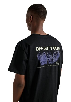 Person mit kurzen Haaren und Creolenohrring trägt ein schwarzes T-Shirt mit dem Text "OFF DUTY GEAR" und einer lila Reifenprofil-Grafik auf der Rückseite.