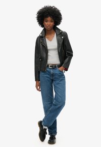 Chaqueta de motociclista de cuero negro con cremalleras plateadas, camiseta gris de cuello en V, vaqueros azul rectos y zapatos negros con suela gruesa.