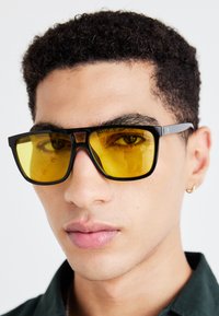 Ochelari de soare negri cu lentile galbene, de formă rectangulară, cu ramă din plastic groasă, având un design minimal și o branduire subtilă pe templa.