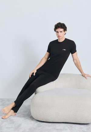 T-shirt preta de algodão com um pequeno logótipo bordado, combinada com calças de lazer pretas correspondentes, usada por um modelo sentado em um sofá claro texturizado.