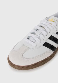 White athletic shoe with black stripes, suede and synthetic material, rounded toe, laces, and textured brown rubber outsole.