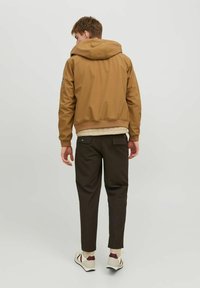 Veste à capuche marron avec des poignets et un ourlet côtelés, portée par-dessus un top crème matelassé, associée à un pantalon marron foncé et des baskets beiges.