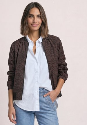 Femme aux cheveux bruns mi-longs portant une veste bomber sombre à motifs sur une chemise blanche et un jean bleu, une main dans la poche.