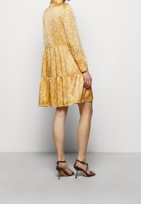 Robe florale jaune à manches longues et jupe à volants, fabriquée en tissu léger. Associée à des sandales à talons hauts noires à lanières.