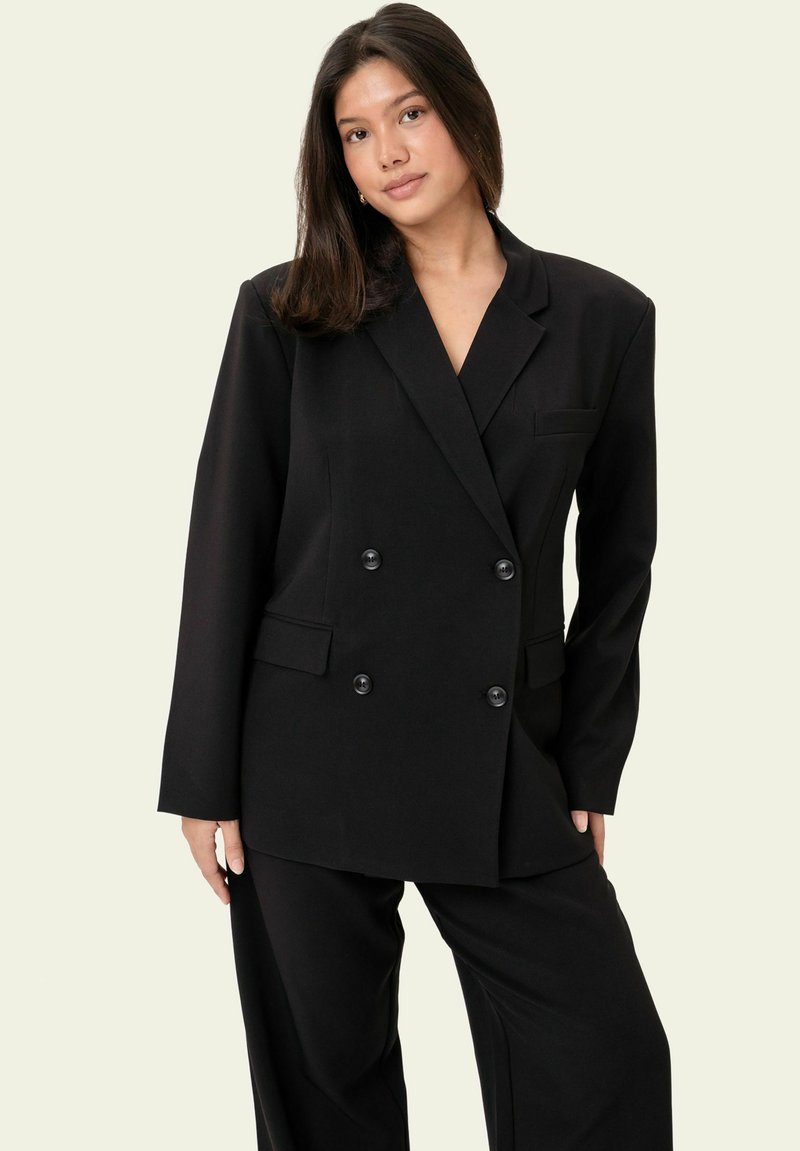 Femme aux cheveux lisses et foncés portant un blazer noir oversized à double boutonnage et un pantalon noir assorti, sur un fond uni.