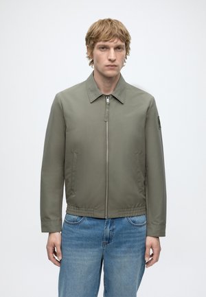 Mand med lysebrunt hår iført en olivengrøn jakke med lynlås og blå jeans, står mod en ensfarvet lys baggrund.